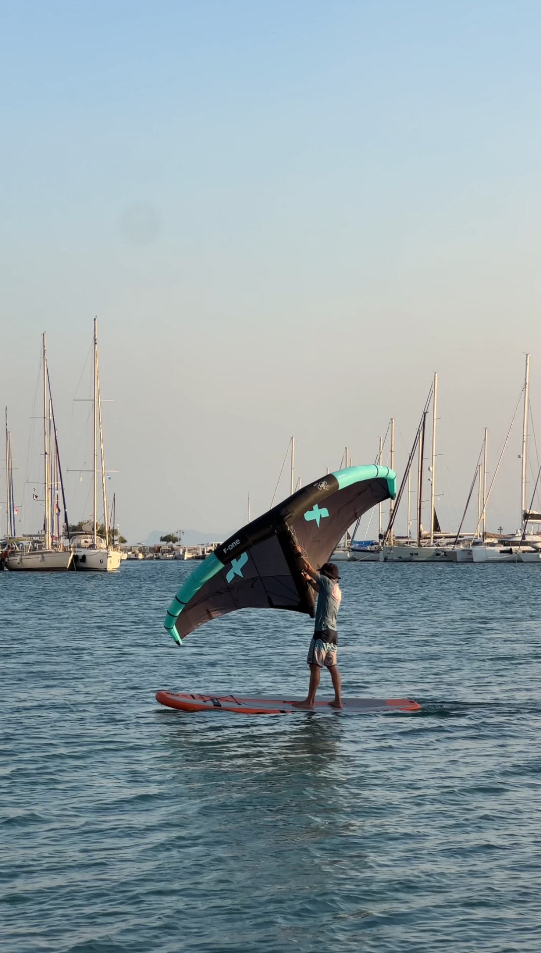 Wing surfer / wing foiler riding a board with a wing in front of moored sailboats