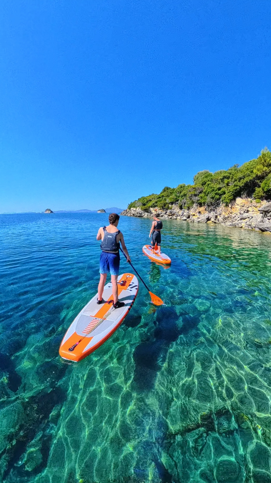 Two paddleboarders gliding over crystal clear Ionian water along a rocky shore