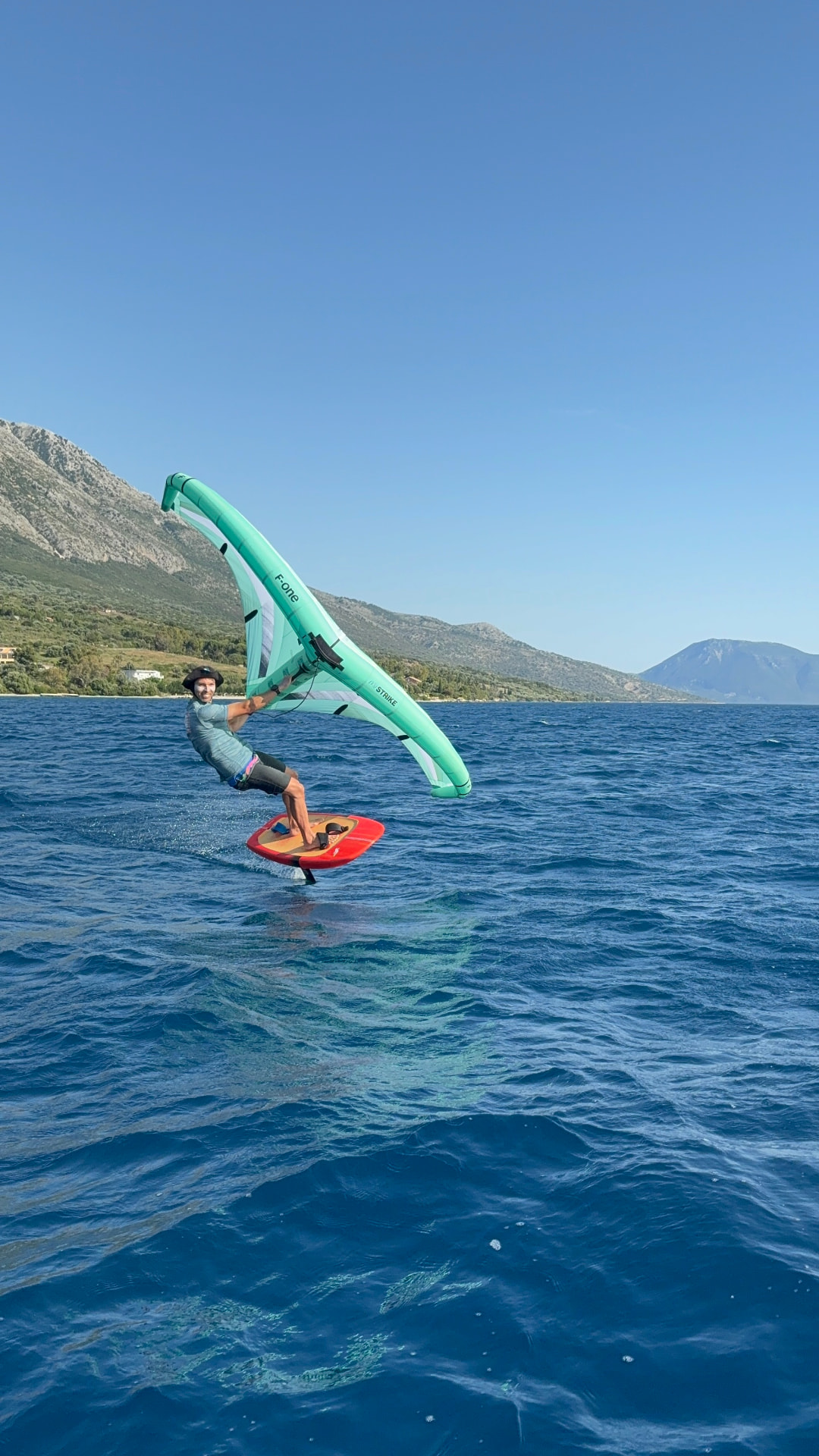 Wing foiler carving with a green wing in front of the mountains near Paleros