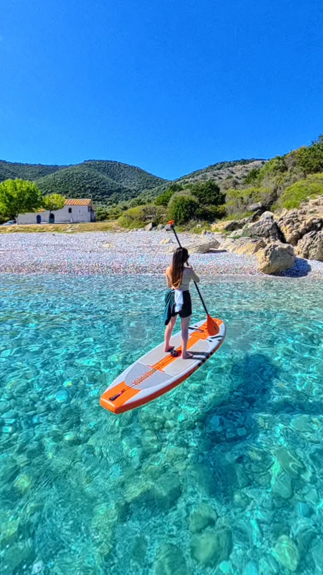 Solo paddleboarder on turquoise water beside a pebble beach and small chapel